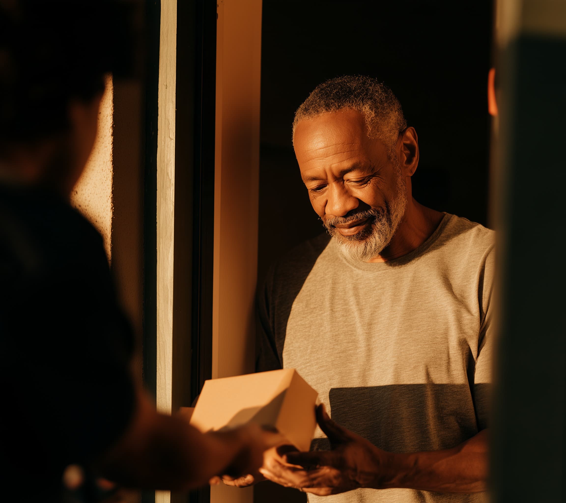 A person in a grey T-shirt at their front door in evening light, smiling as they take a small box